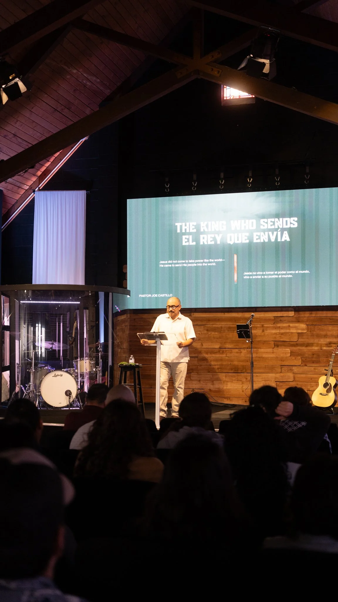 A man speaking at a church or conference stage with a large screen behind him displaying text in both English and Spanish about Jesus being the king who sends the Messiah. The stage has musical instruments including drums and a guitar, and a small audience is seated in front.