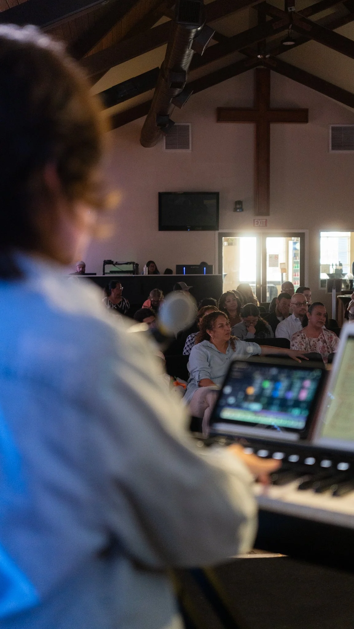 A person in the foreground is blurred and appears to be playing a keyboard or piano, while a congregation listens attentively inside a church with a large cross on the wall and sunlight coming from the open door in the background.