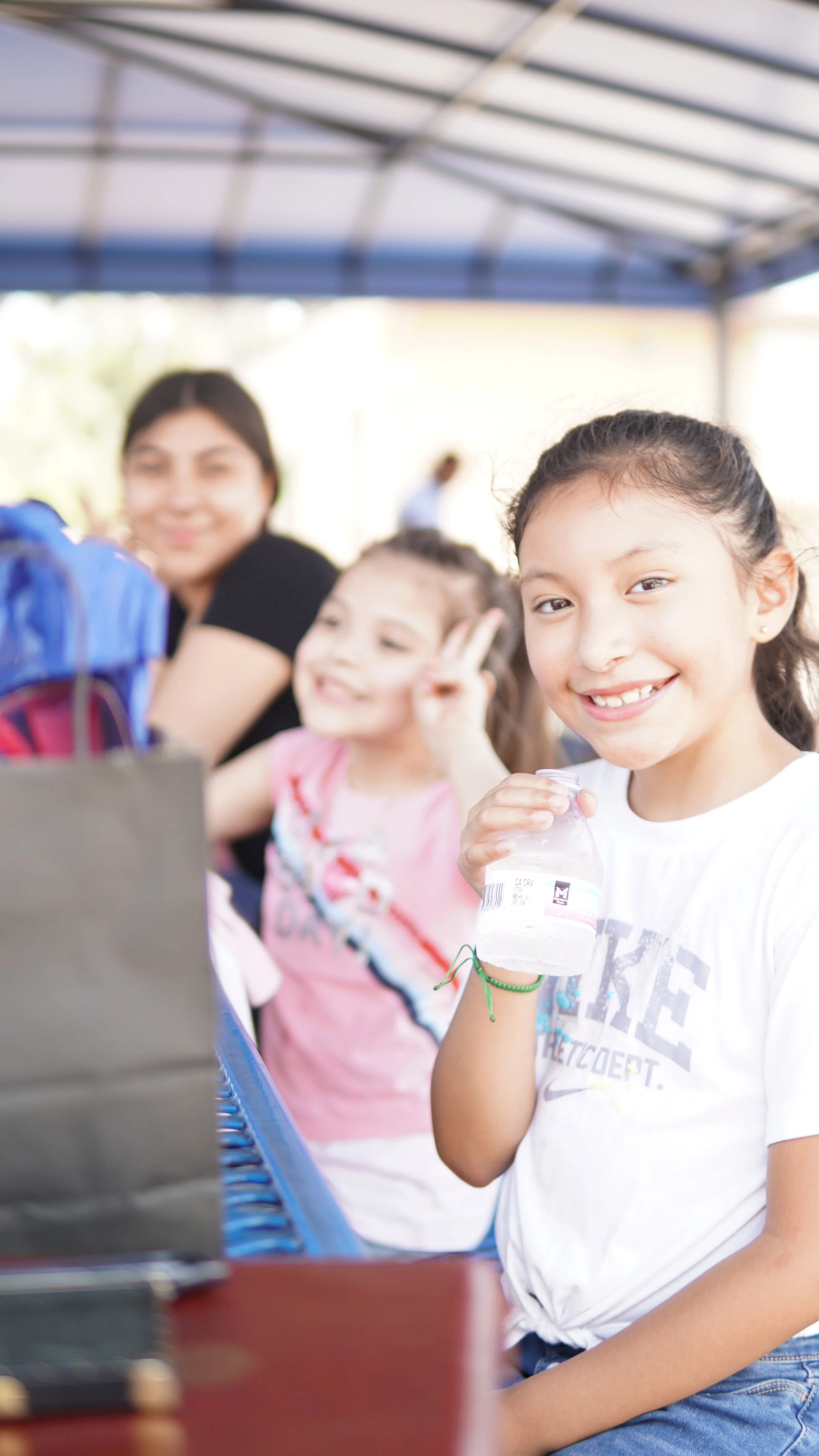 Smiling young girl in a white t-shirt holding a water bottle at an outdoor event with other children and a woman in the background.