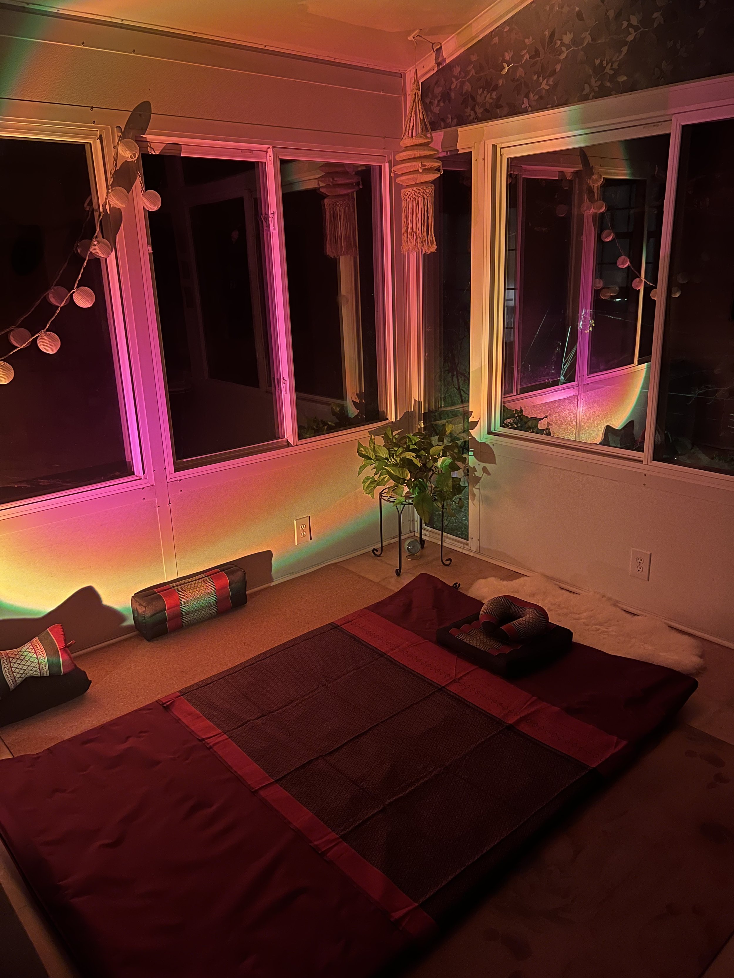 Room interior with a yoga or meditation setup, featuring a mat, cushions, decorative lights, and hanging macrame, illuminated by colorful lighting.