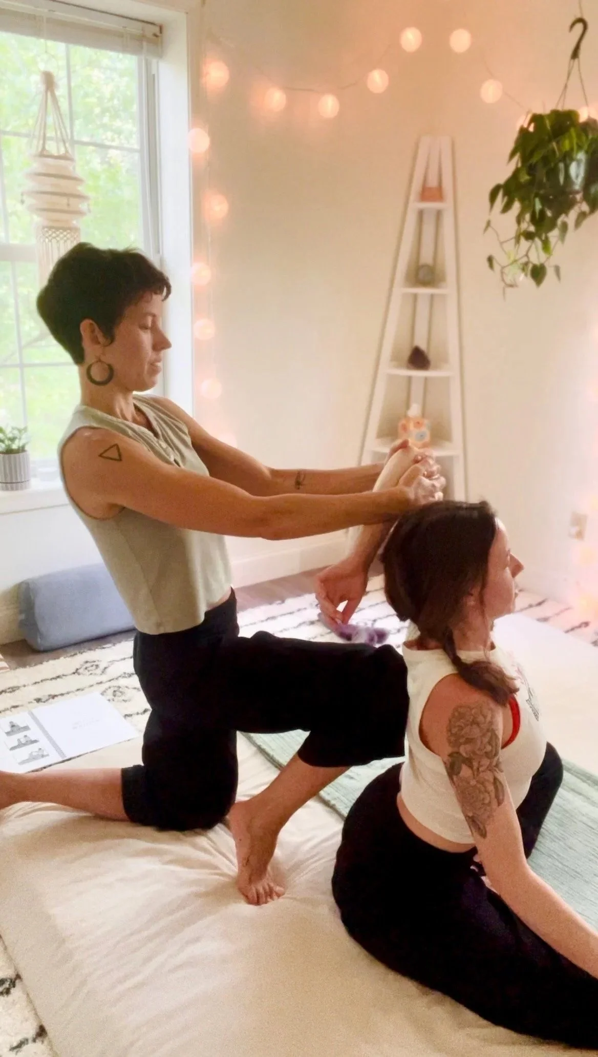 Two people practicing yoga on a porch, with one person assisting the other in a seated side stretch. String lights hang in the background.