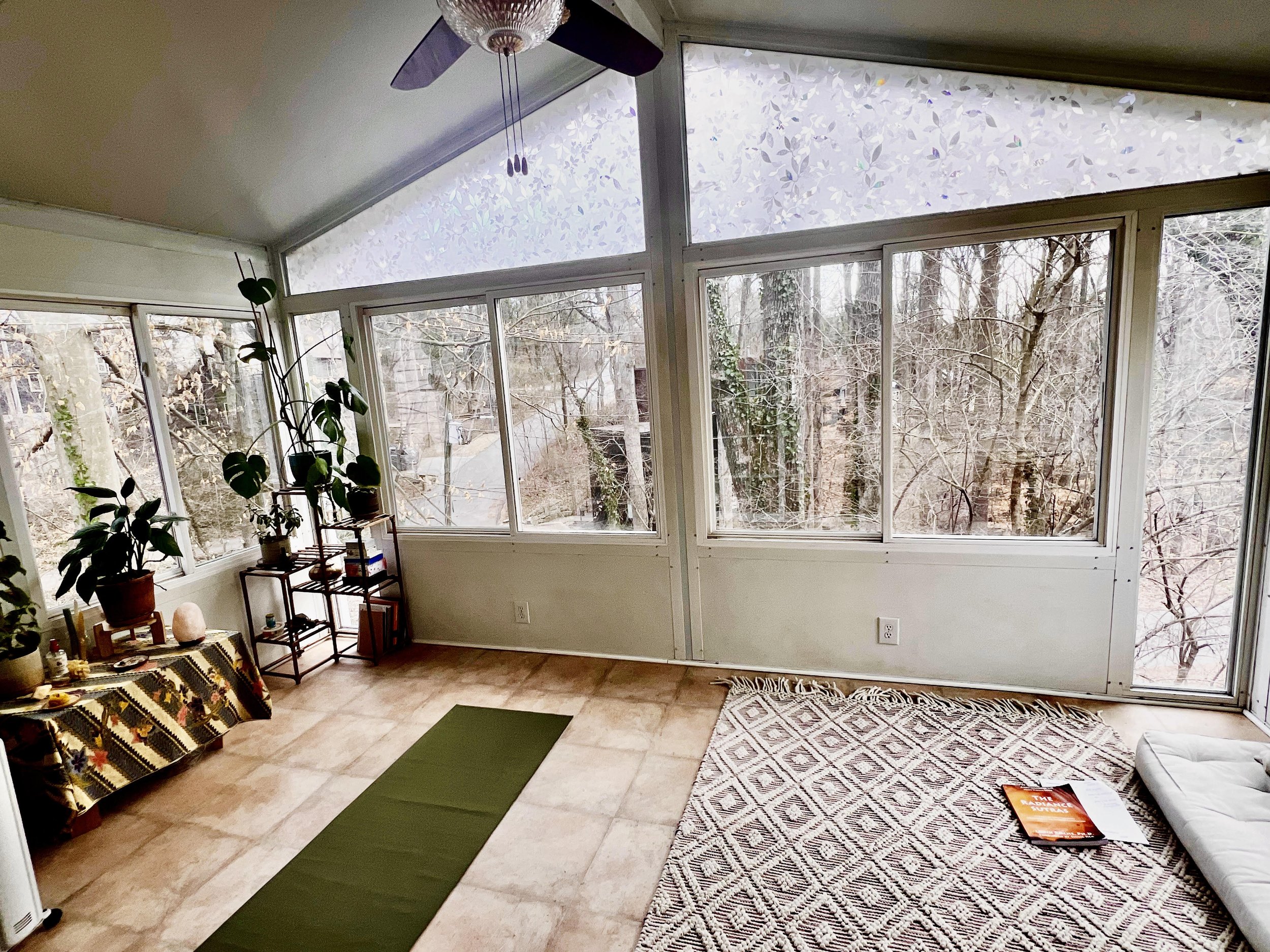 Sunroom with large windows, plants, a yoga mat, patterned rug, and decorative table with various items. Ceiling fan above, trees visible outside.