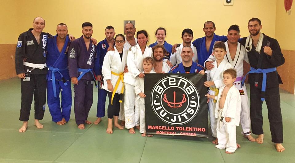 Group of Judo practitioners, including children and adults, standing on a dojo mat with some wearing gi uniforms and others in casual or team gear, holding a banner for Arena Jiu Jitsu with Marcello Tolentino from Montreal, Canada.