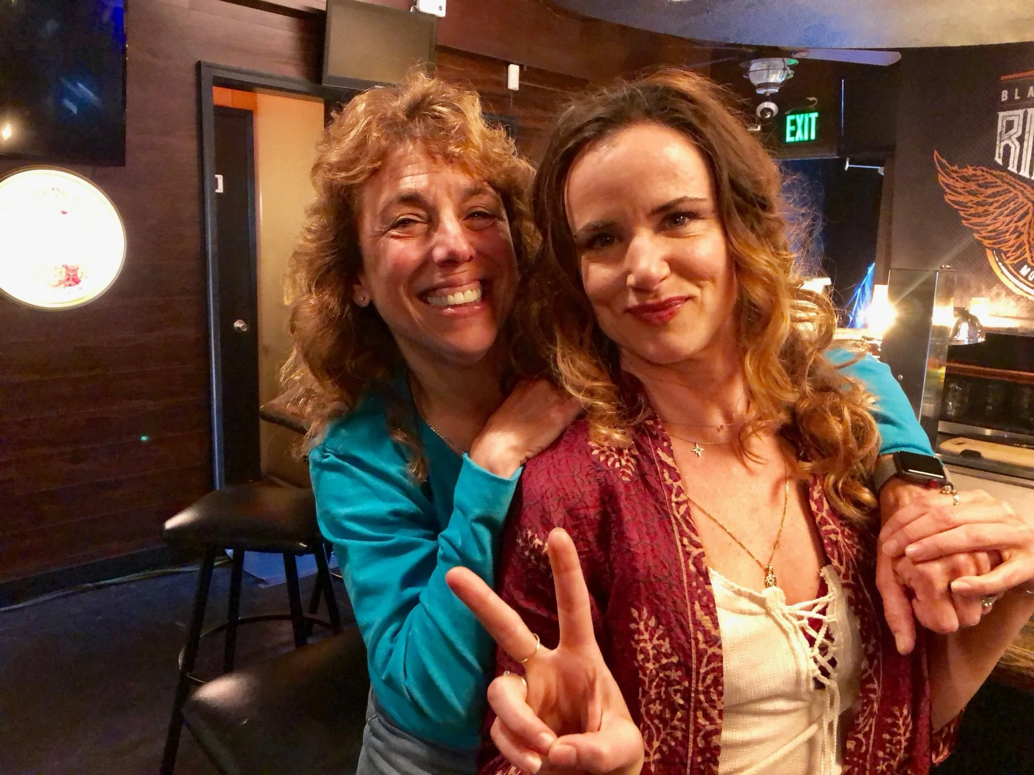 Two women smiling and posing for a photo in a dimly-lit room, one holding up a peace sign. They are surrounded by bar stools and a wooden interior, with a wall decoration in the background.