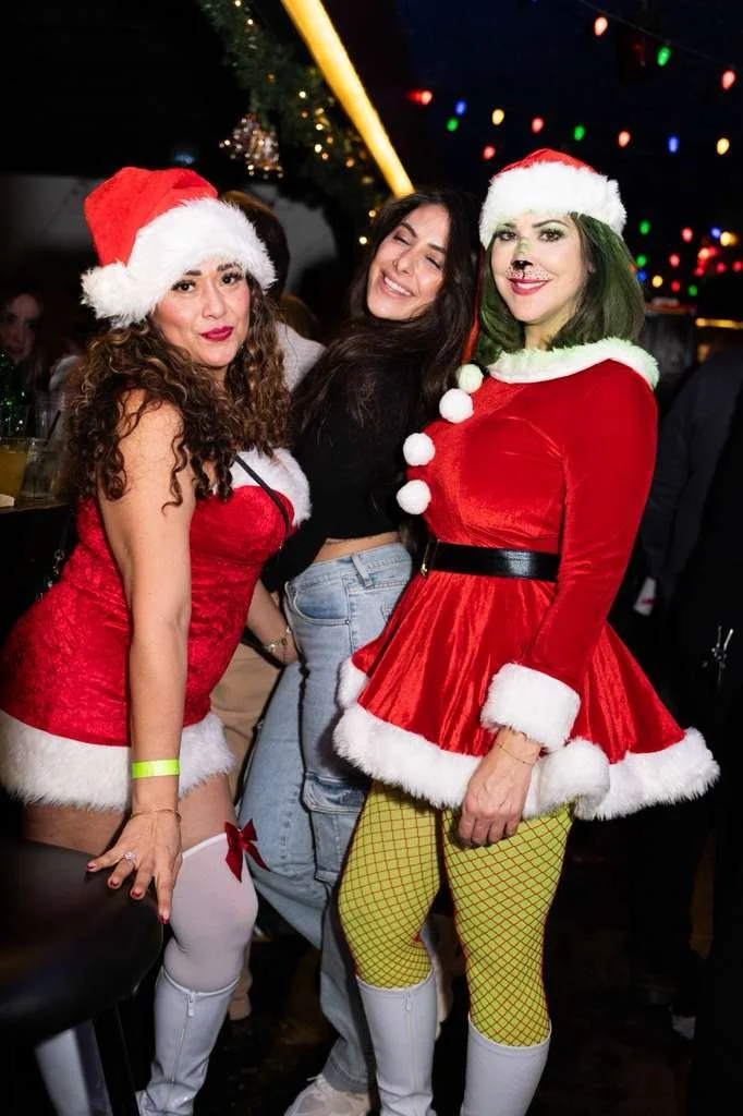Three women wearing Christmas costumes and Santa hats at a holiday party, with colorful Christmas lights in the background.