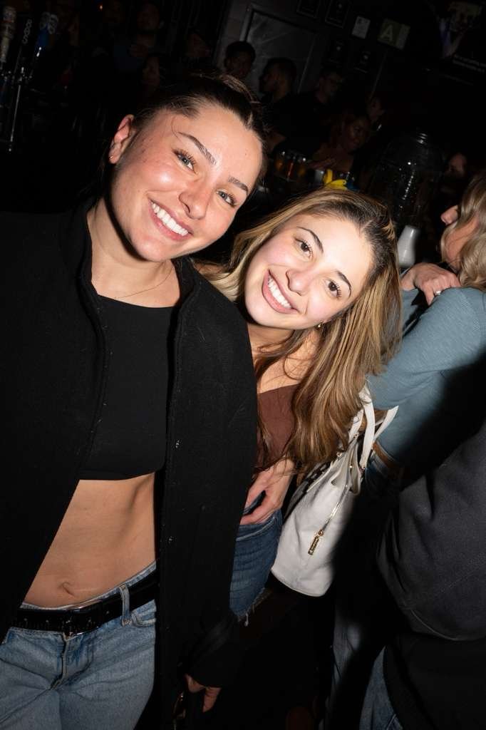 Two young women smiling and posing for a photo in a crowded indoor setting, possibly a bar or club, with others in the background.