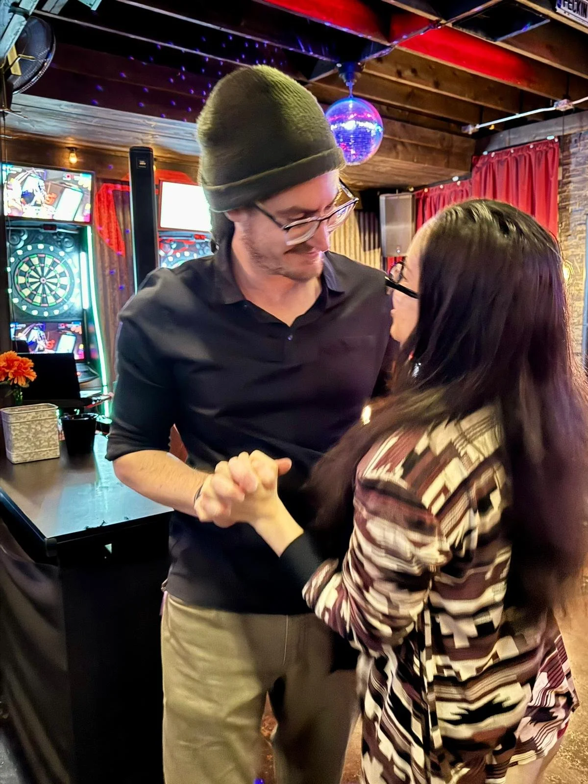 A man and woman are dancing and smiling in a dimly-lit bar or club with dartboards and colorful lights in the background.