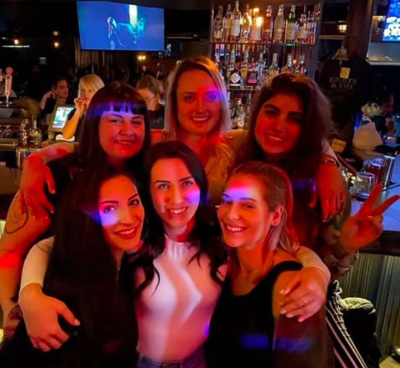 Group of six women smiling and posing together in a bar with colorful lights.