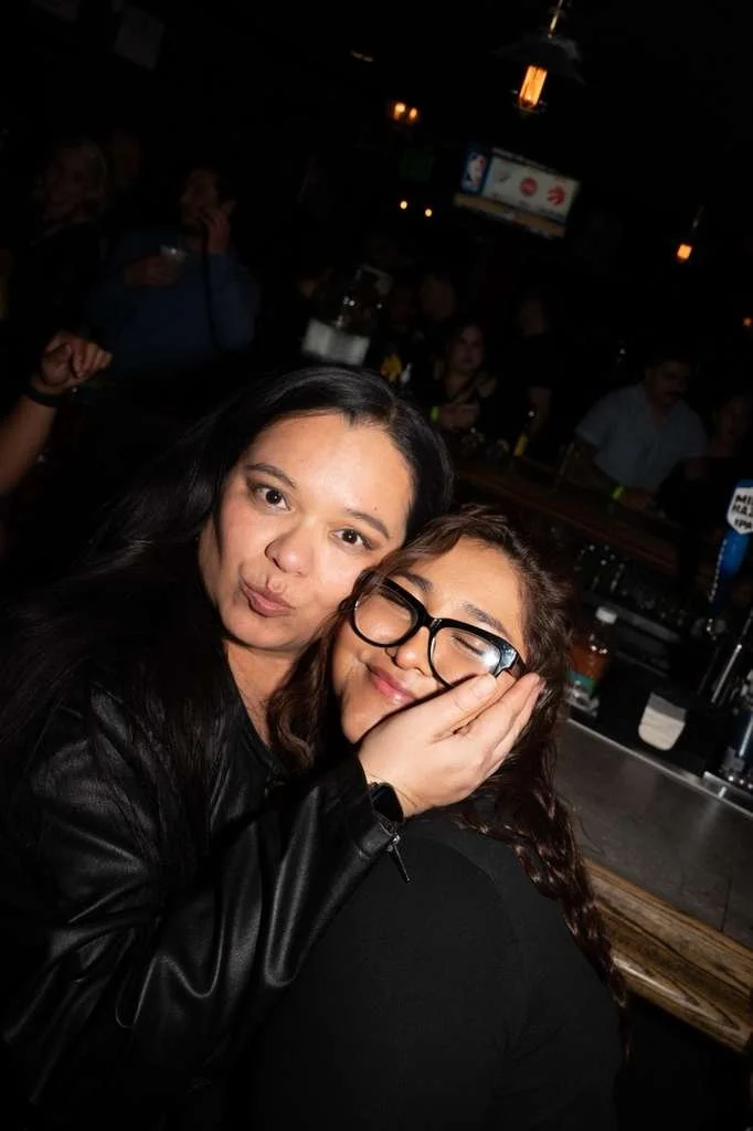 Two women hugging in a dimly lit bar or club, with one woman making a playful expression and the other smiling with glasses, surrounded by other people and bar equipment in the background.