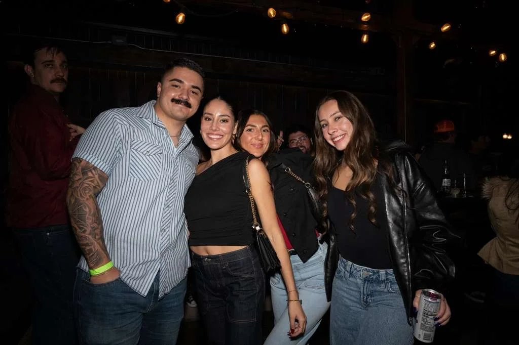 Group of five friends smiling and standing together in a dimly lit bar or club, some holding drinks.