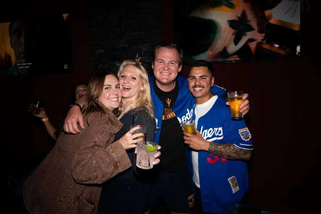 Four friends smiling and holding drinks in a bar with dark walls and artwork in the background, celebrating together.