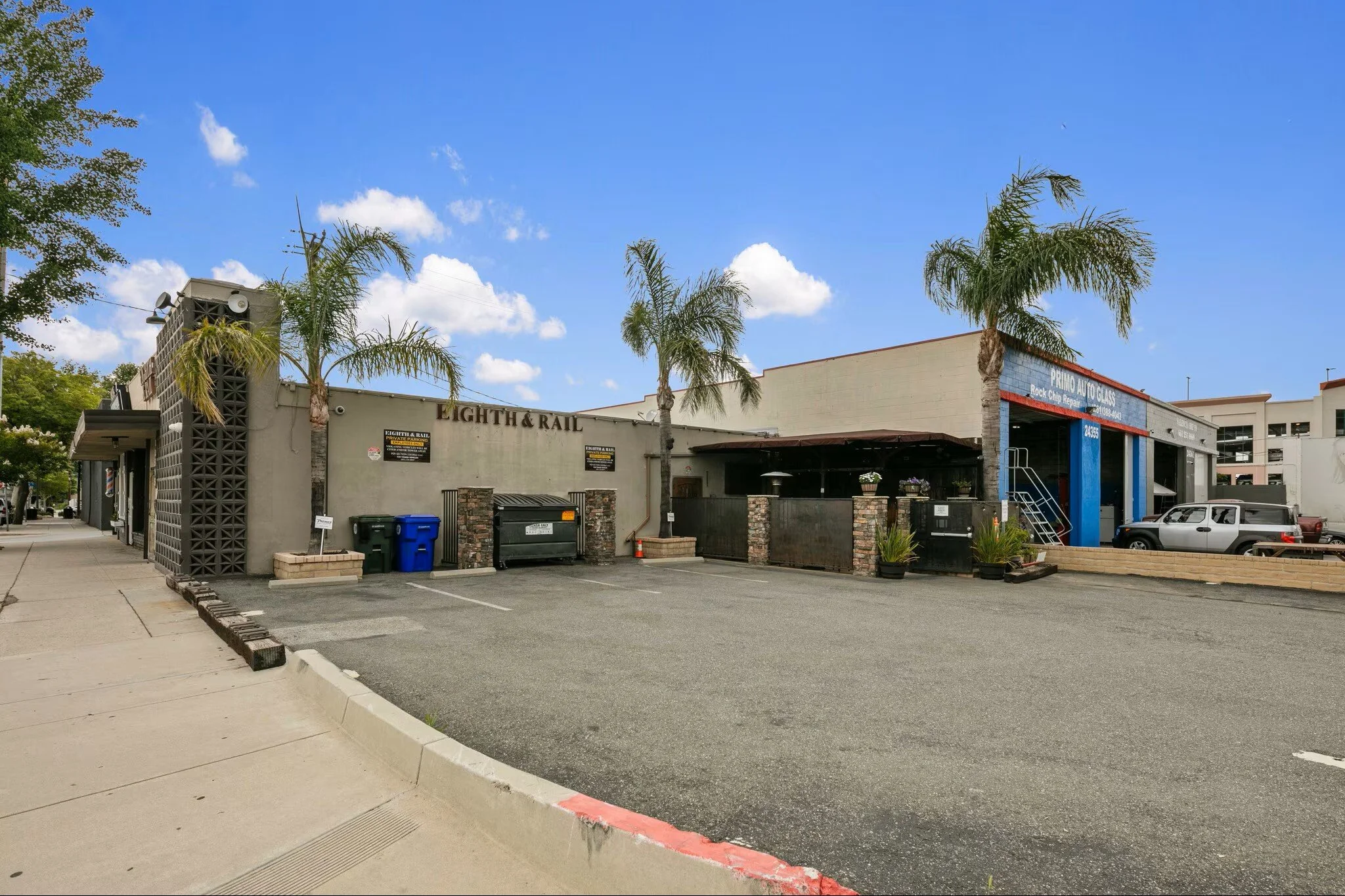 A commercial building with two businesses, Eighth & Rail and Primo Auto Glass, situated on a street corner with a parking lot, palm trees, and a clear blue sky.