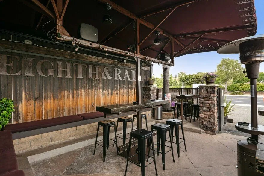 Outdoor seating area of a restaurant named 'DIGHT & RAL' with a high table, bar stools, a bench, and a heater, under a large umbrella, with trees and a road visible in the background.