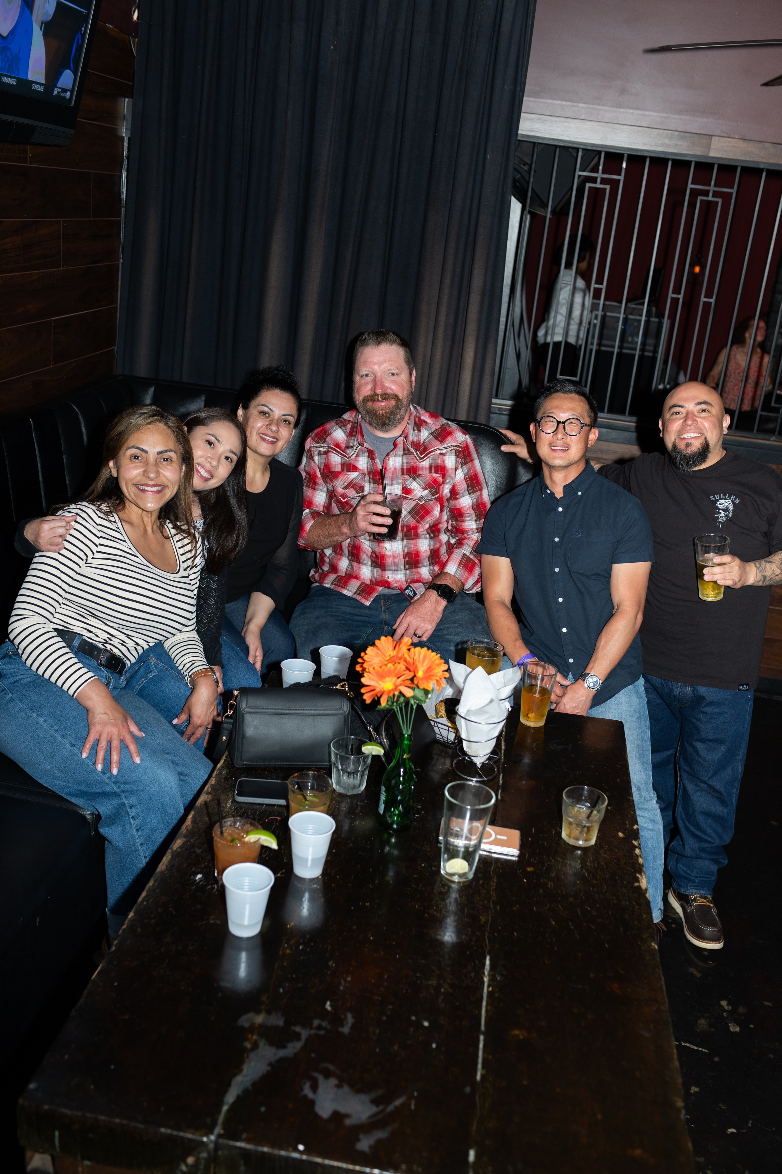 Group of six diverse friends sitting together at a bar or restaurant, smiling and holding drinks, with a black leather booth, a dark wooden table with flowers, and a dark curtain in the background.