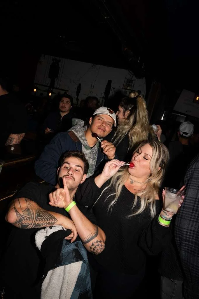 Group of four young adults at a bar or nightclub, posing for a photo with drinks, some making playful gestures, and others enjoying the moment.