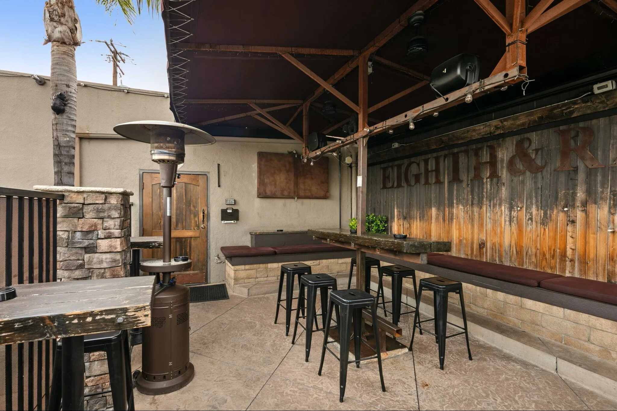 Empty outdoor patio area with a wooden bar area, black bar stools, a heater, and a wall with the words 'EIGHTH & R' in a casual restaurant or bar setting.