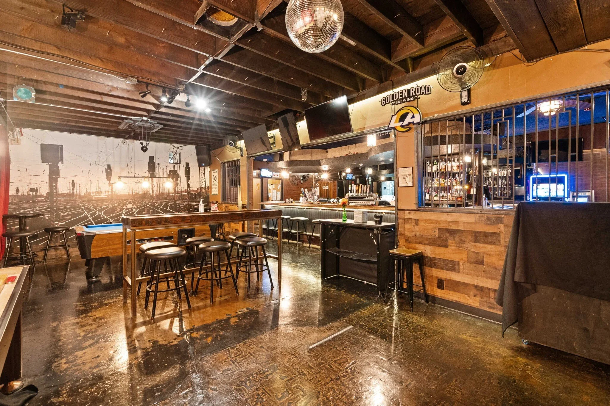 Interior of a bar or pub with high tables and stools, a large mural of train tracks at sunset on the wall, four television screens, a bar counter with taps, and a neon sign that reads 'LA.' The ceiling is wooden with exposed beams, and there is a disco ball hanging from it.