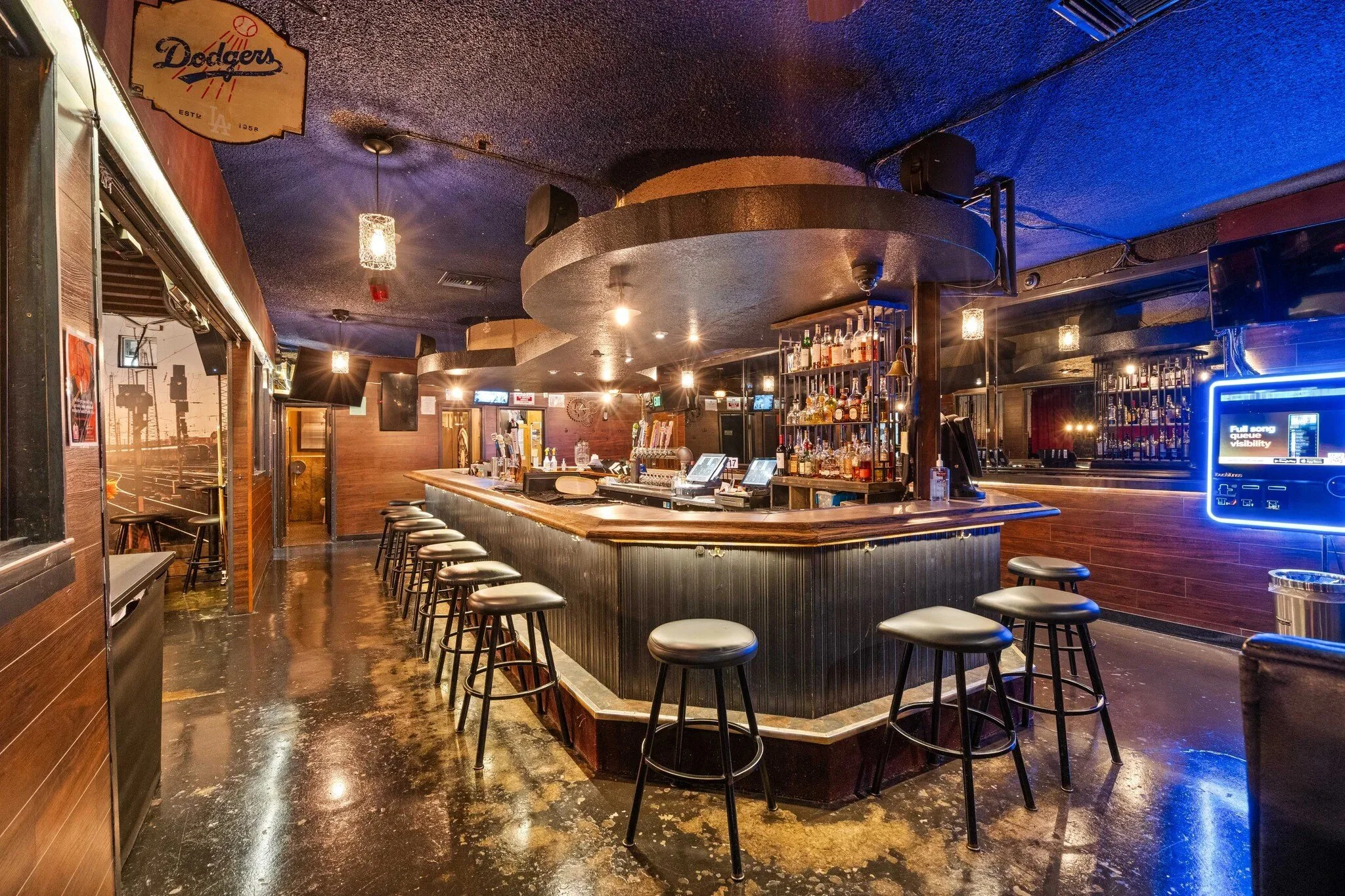 Empty bar with black stools around a curved wooden counter, bottles of alcohol on shelves, and blue lighting accents.