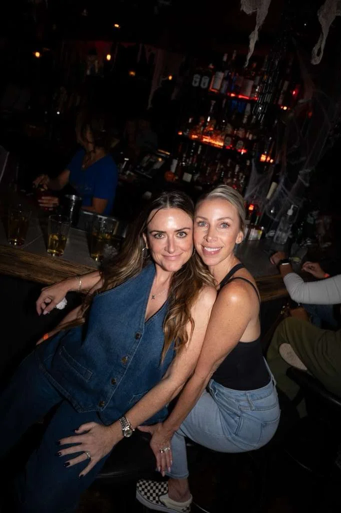 Two smiling women sitting at a bar in a dimly lit restaurant or bar with a display of bottles behind them.