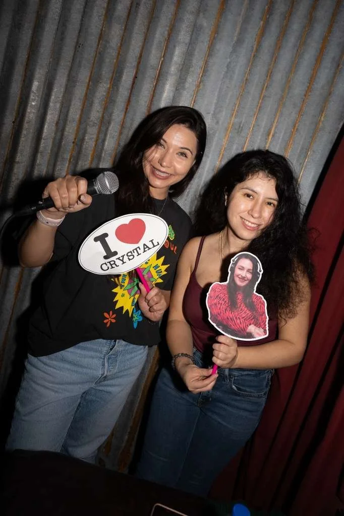 Two women standing together smiling, one holding a microphone and a speech bubble sign that says 'I ♥ CRYSTAL,' the other holding a cutout photo of a woman in a red dress. They are in front of a striped curtain backdrop.