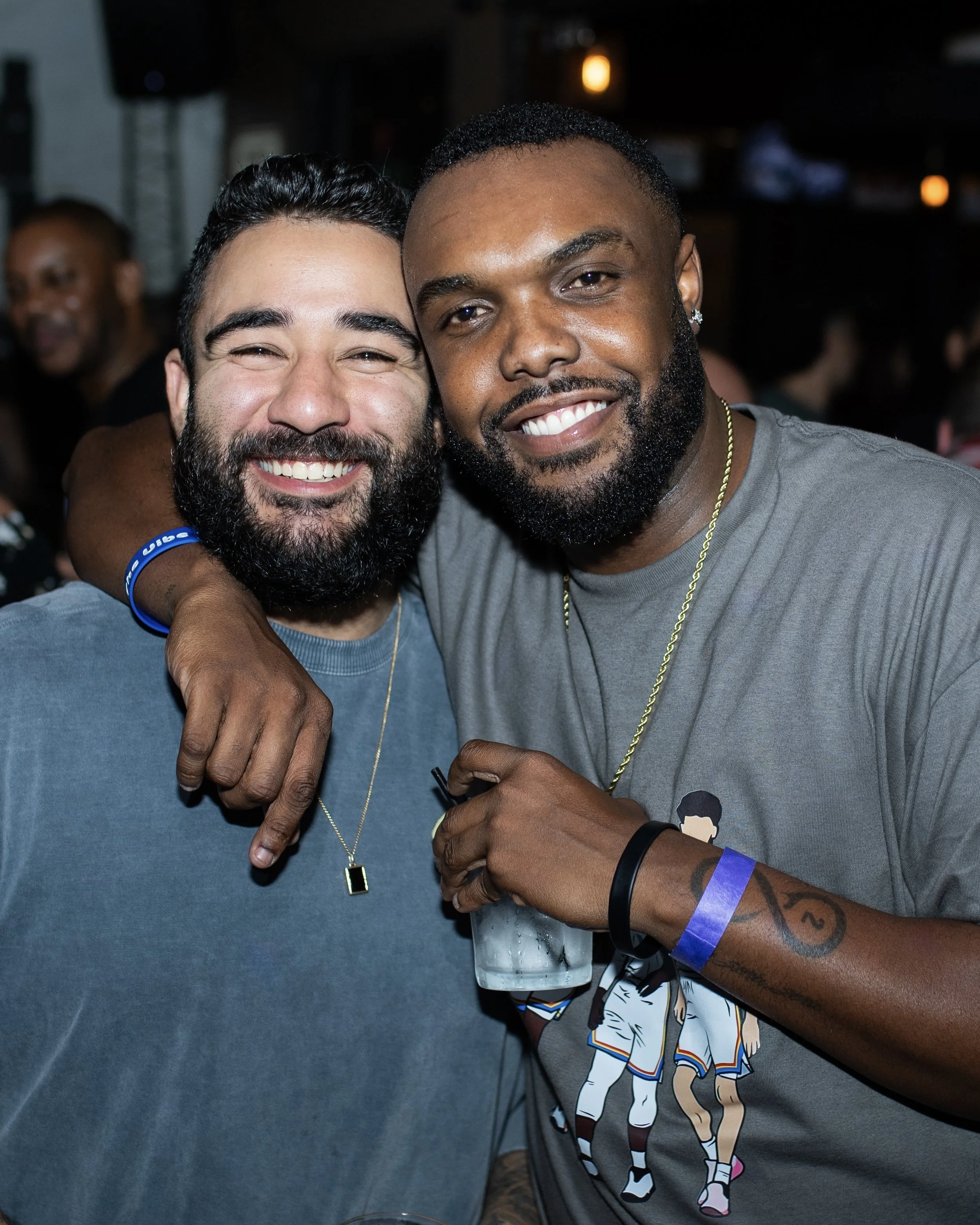 Two men smiling and hugging at a social event, one with a beard and wearing a blue T-shirt, the other with a beard and wearing a gray T-shirt with a basketball graphic.