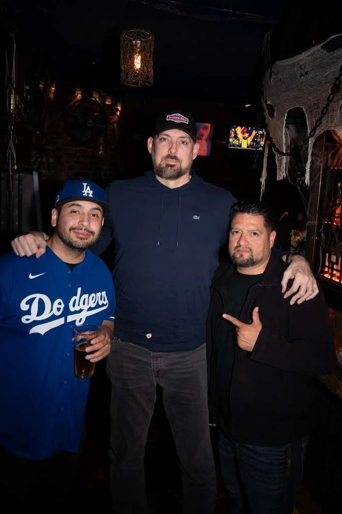 Three men posing together in a dimly lit bar or pub, with one man wearing a Dodgers jersey and holding a drink, another in a black jacket pointing at the middle man, and the middle man wearing a dark hoodie and a baseball cap.