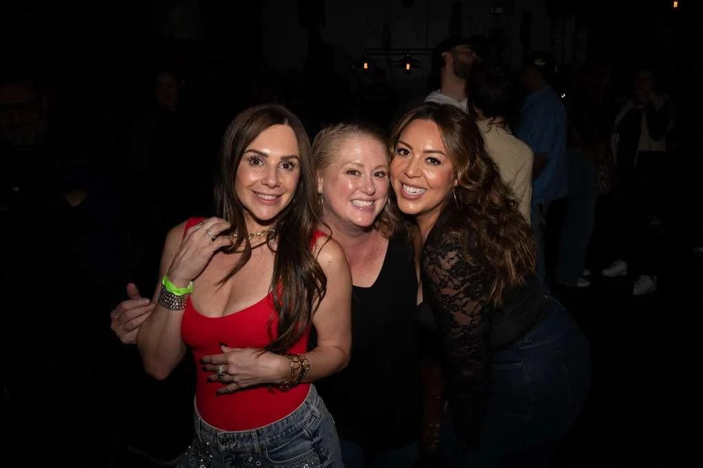 Three women smiling and posing together at a nighttime social event, with other people visible in the background.