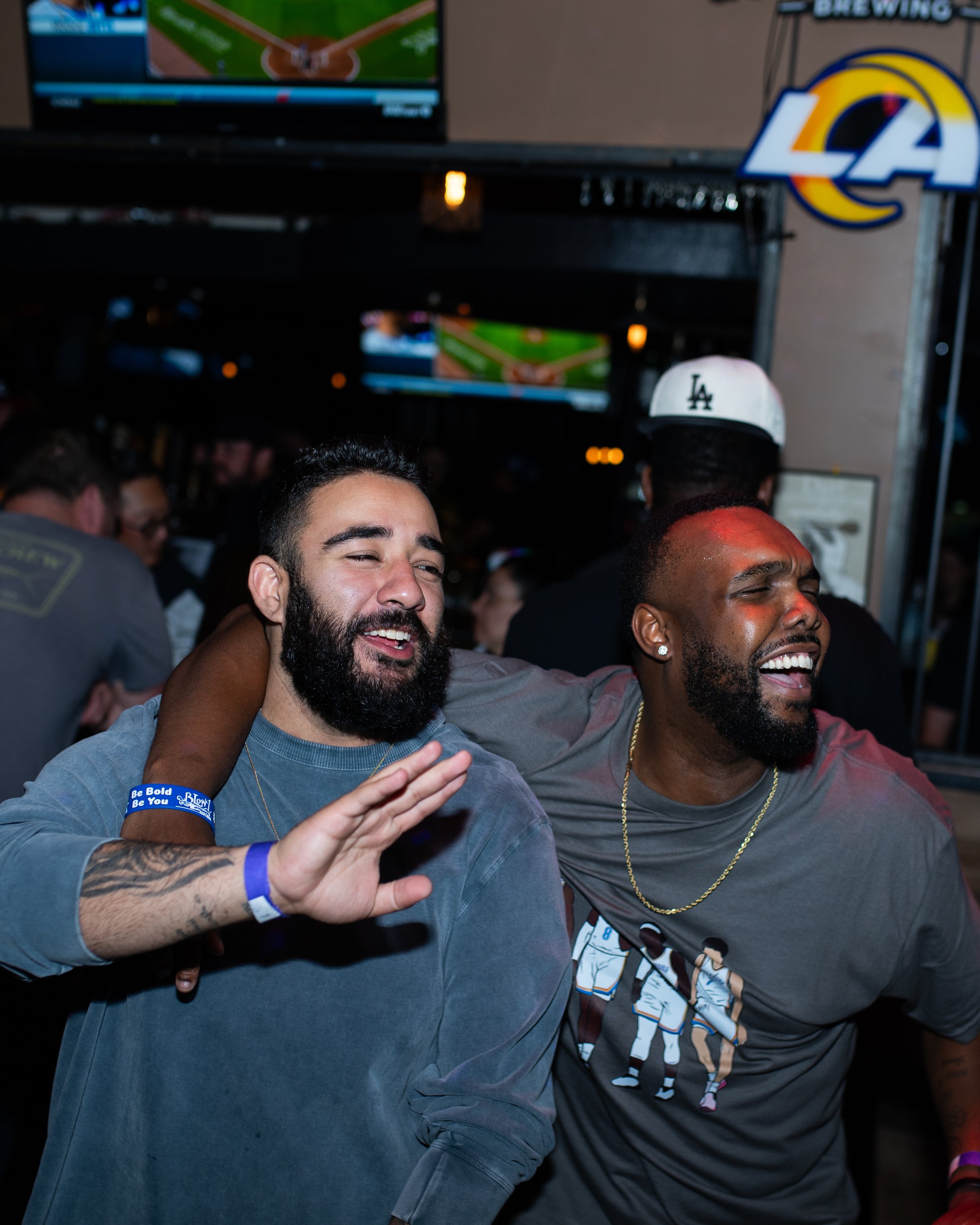 Two men celebrating and smiling at a sports bar or pub, with one wearing a gray shirt with a graphic of basketball players and the other wearing a gray shirt, informally enjoying themselves.