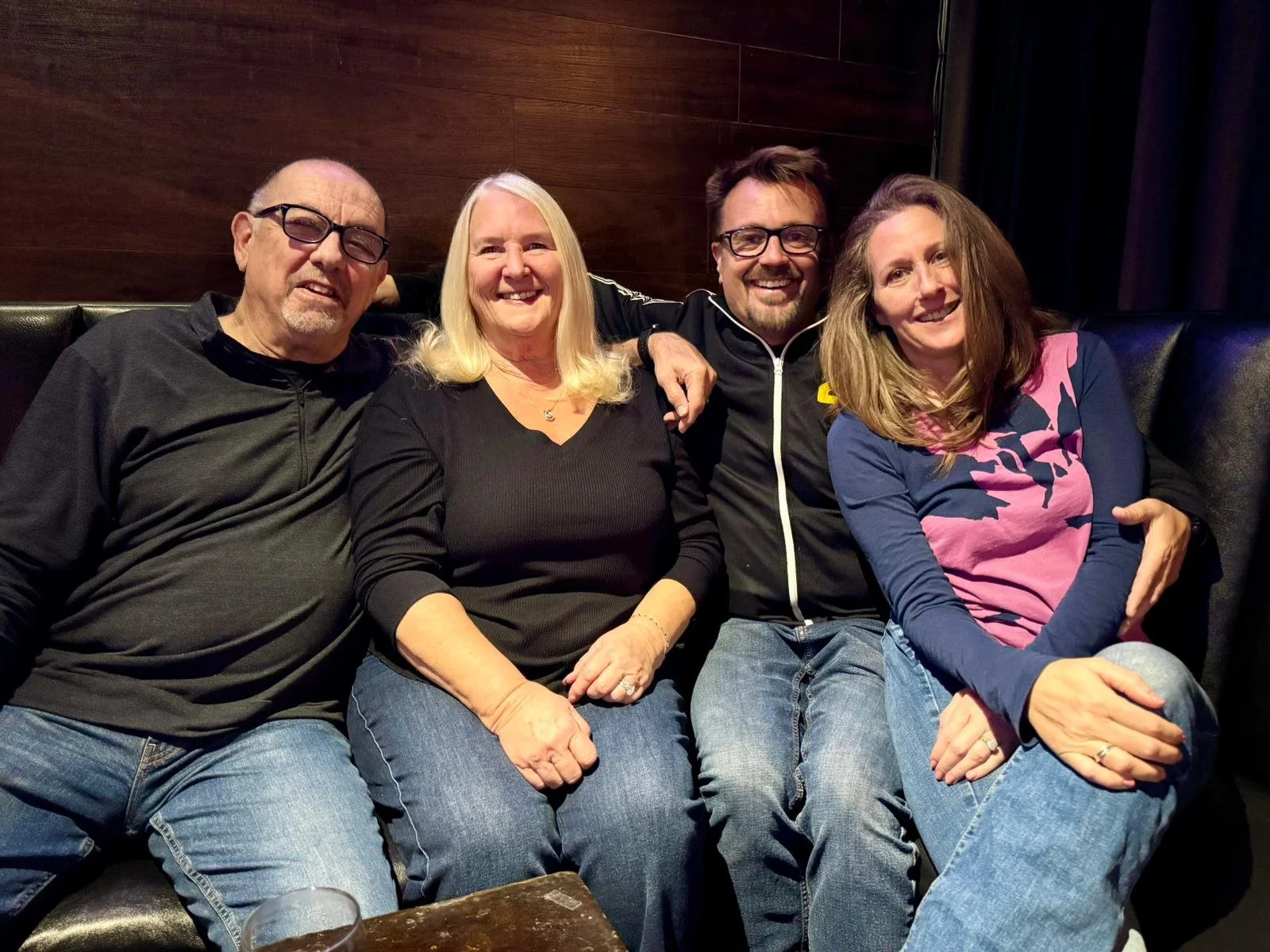 Four people sitting on a black leather couch in a dimly lit room, smiling and posing for the photo. From left to right: an older man with glasses and a black shirt, a woman with blonde hair and a black top, a man with glasses and dark clothing, and a
