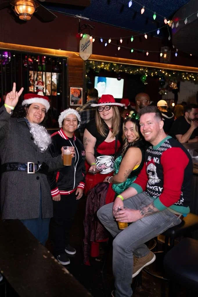 Group of people celebrating Christmas at a bar, wearing holiday hats and sweaters, with Christmas decorations and colorful string lights in the background.