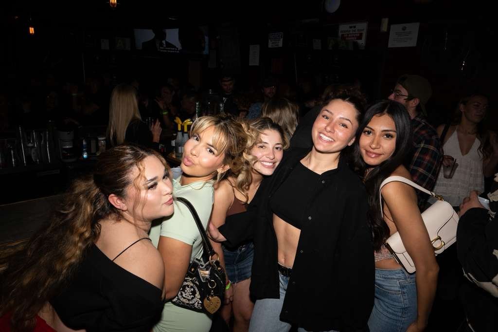 Group of five young women posing in a bar or nightclub with dim lighting and other people in the background.