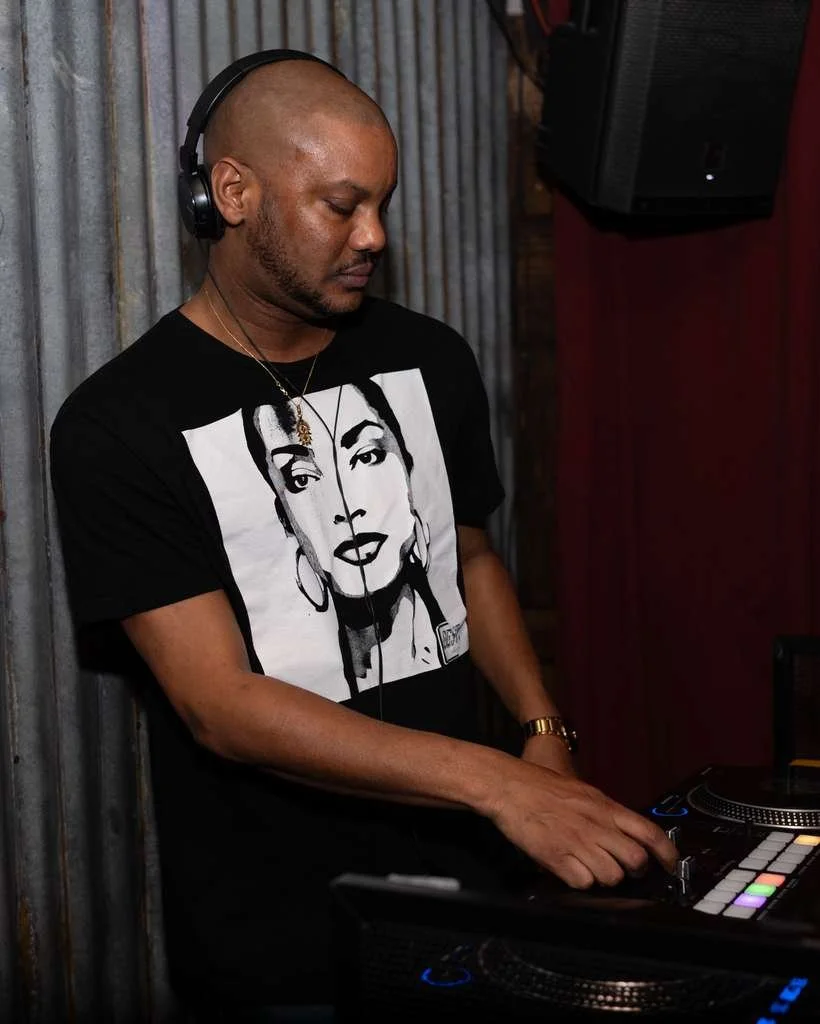 A man with a shaved head wearing headphones and a black T-shirt with an illustrated portrait of a woman, operating DJ equipment in a dimly lit room with corrugated metal wall.