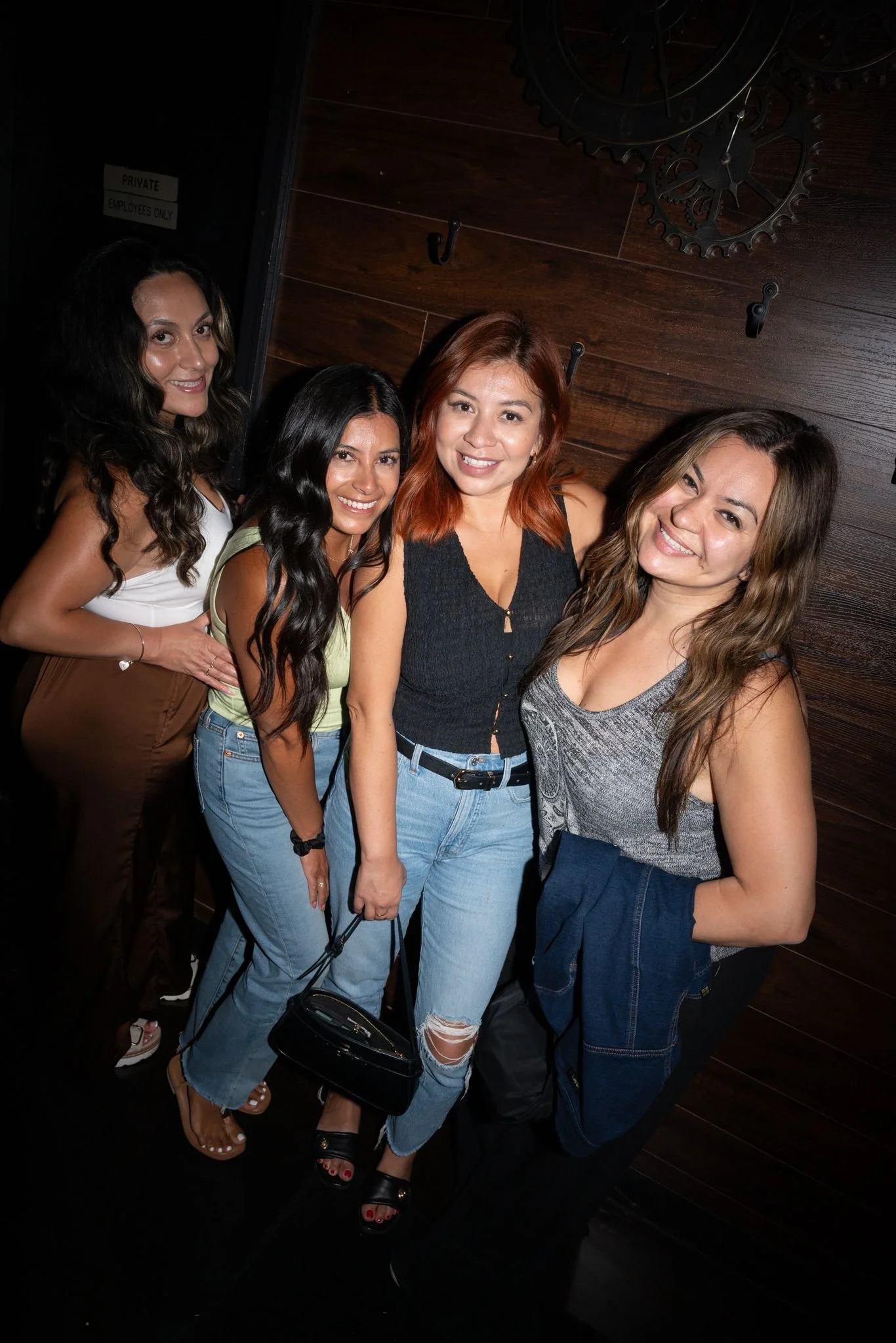 Group of five women smiling and posing together at a social event, standing in front of a dark wooden wall with metal gears decor.