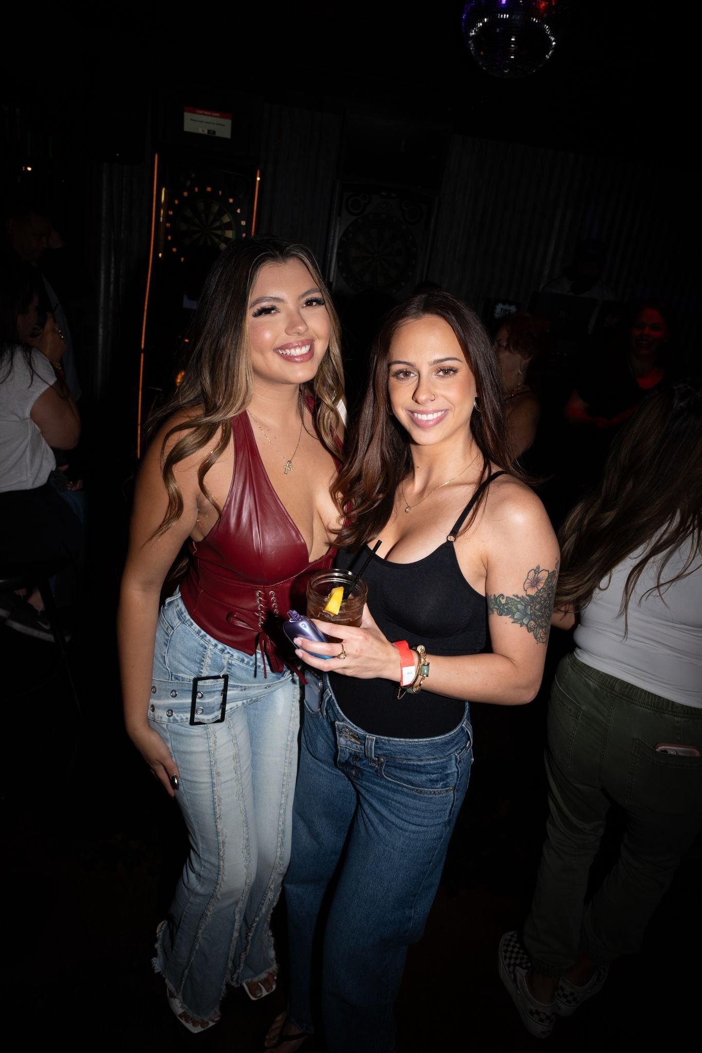Two young women smiling at a dark indoor bar, one holding a drink with a lemon wedge, surrounded by other people.