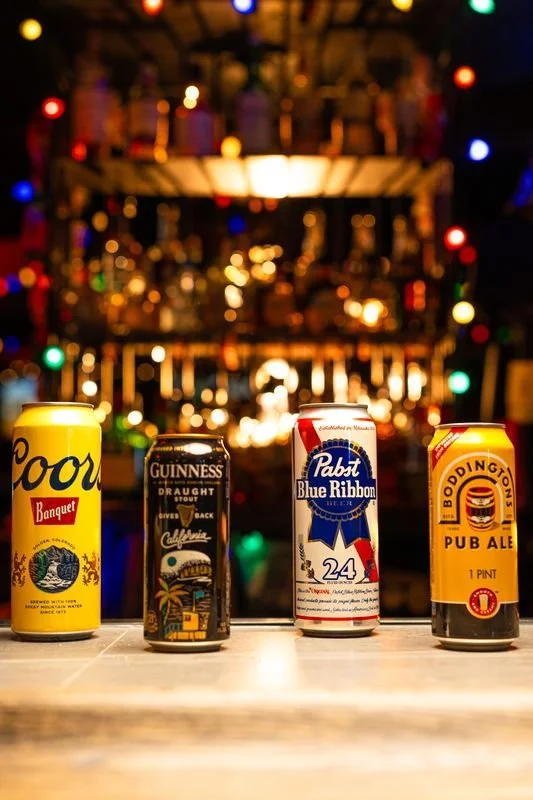 Four cans of beer and stout on a bar counter with colorful blurred lights in the background.