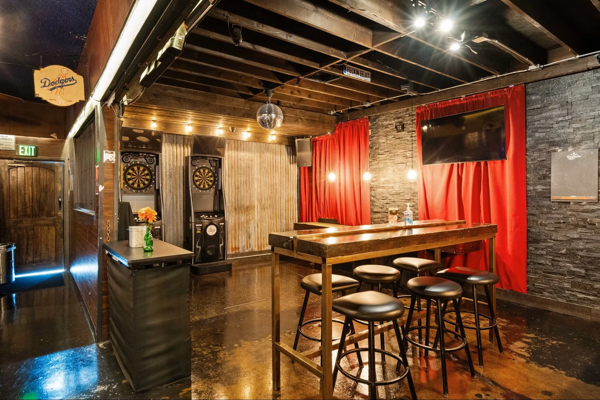 Bar or game room with dartboards, a tall wooden table, stools, black stone wall with red curtains, and a TV.