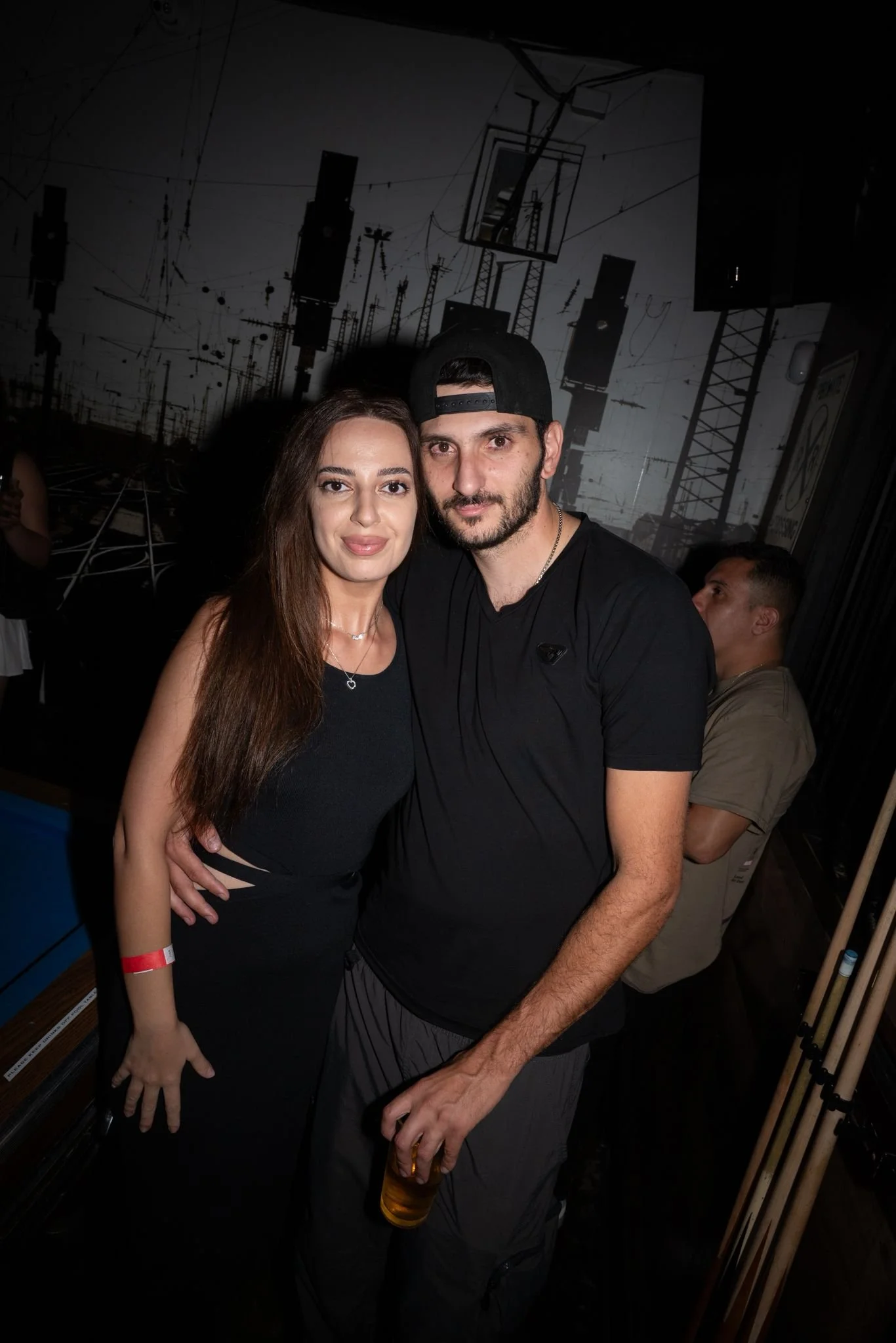 A man and woman posing together at a social event. The woman has long brown hair, wearing a black dress, and the man is wearing a black t-shirt and a black baseball cap, holding a glass of beer.