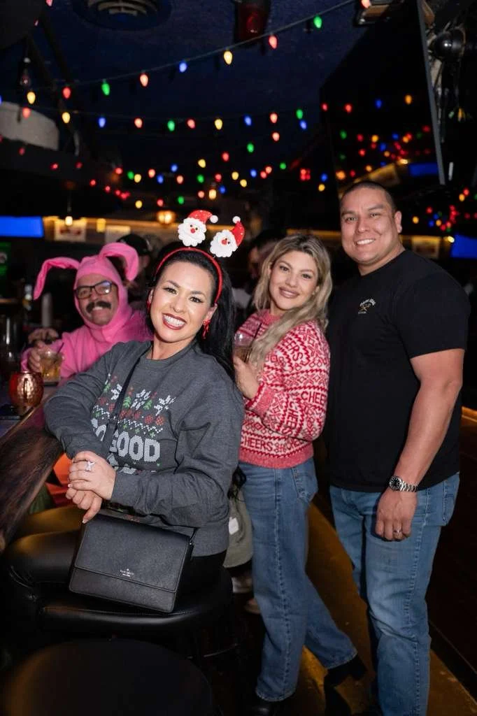People celebrating Christmas at a bar, wearing festive sweaters and headbands, with Christmas decorations and colorful string lights overhead.