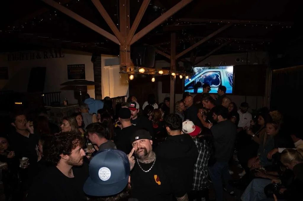 Crowded indoor party with people socializing and watching a large screen showing a futuristic vehicle, dim lighting, and a chandelier with multiple bulbs.
