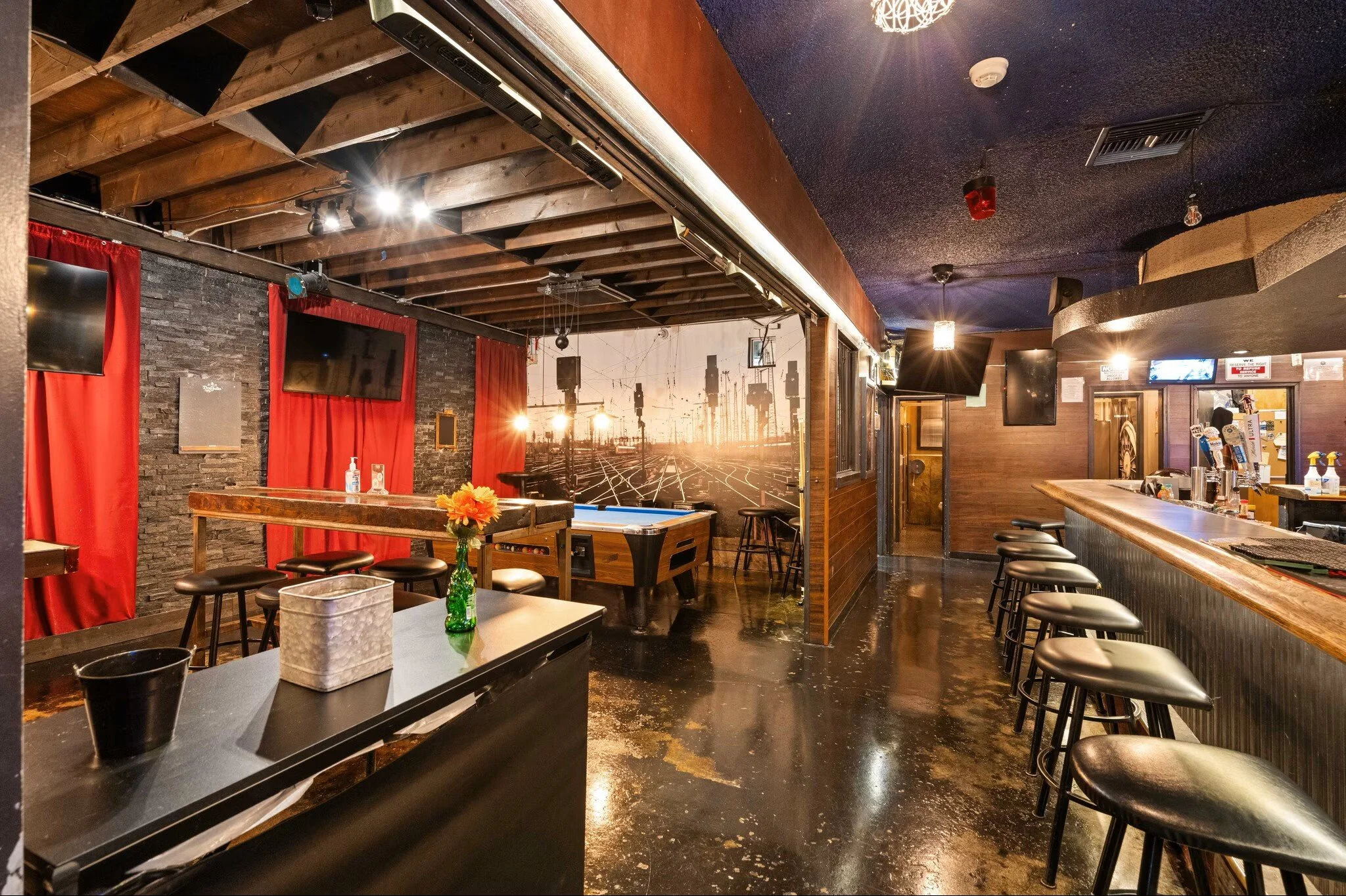 Interior of a bar with black-painted floors, wooden ceiling beams, bar counter with stools, red curtains, and a mural of train tracks and city Skyline in the back.