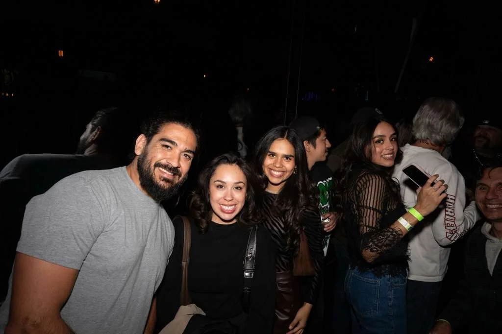 Group of five people smiling at a social event, with others in the background, dark lighting.