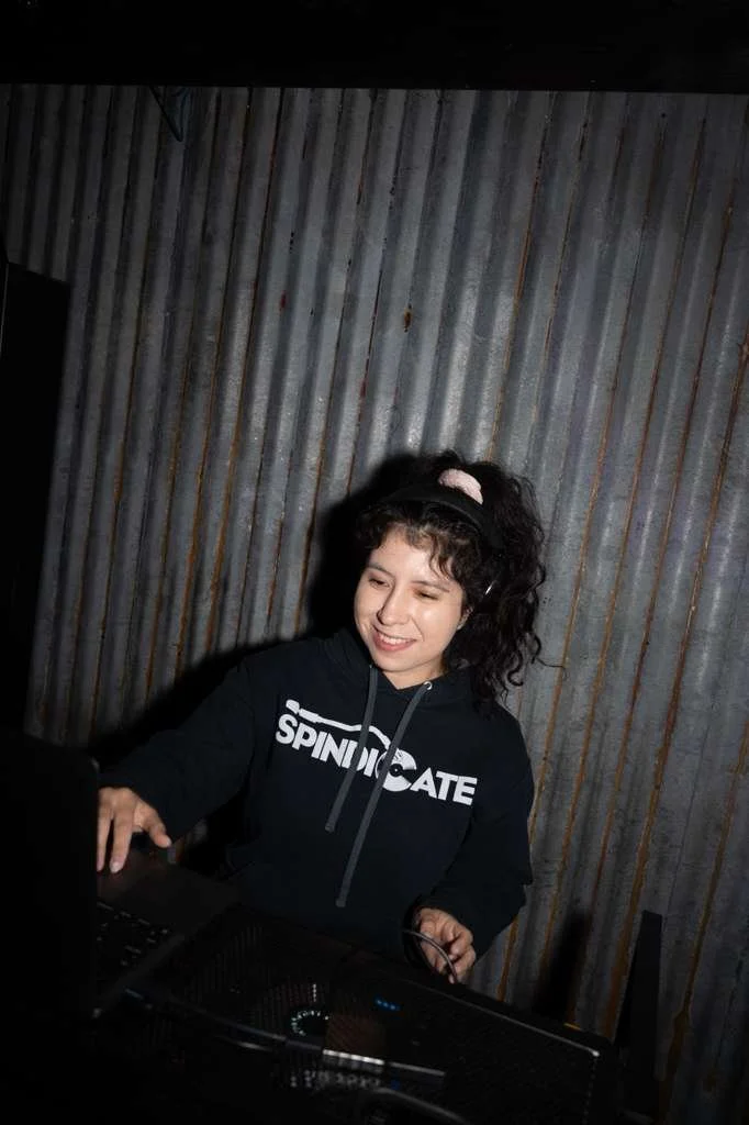 A young woman with curly dark hair and a pink scrunchie, wearing a black Hoodie that says "SPINDICATE," is smiling and looking at a laptop screen in a dimly lit room with a metallic, corrugated wall behind her.
