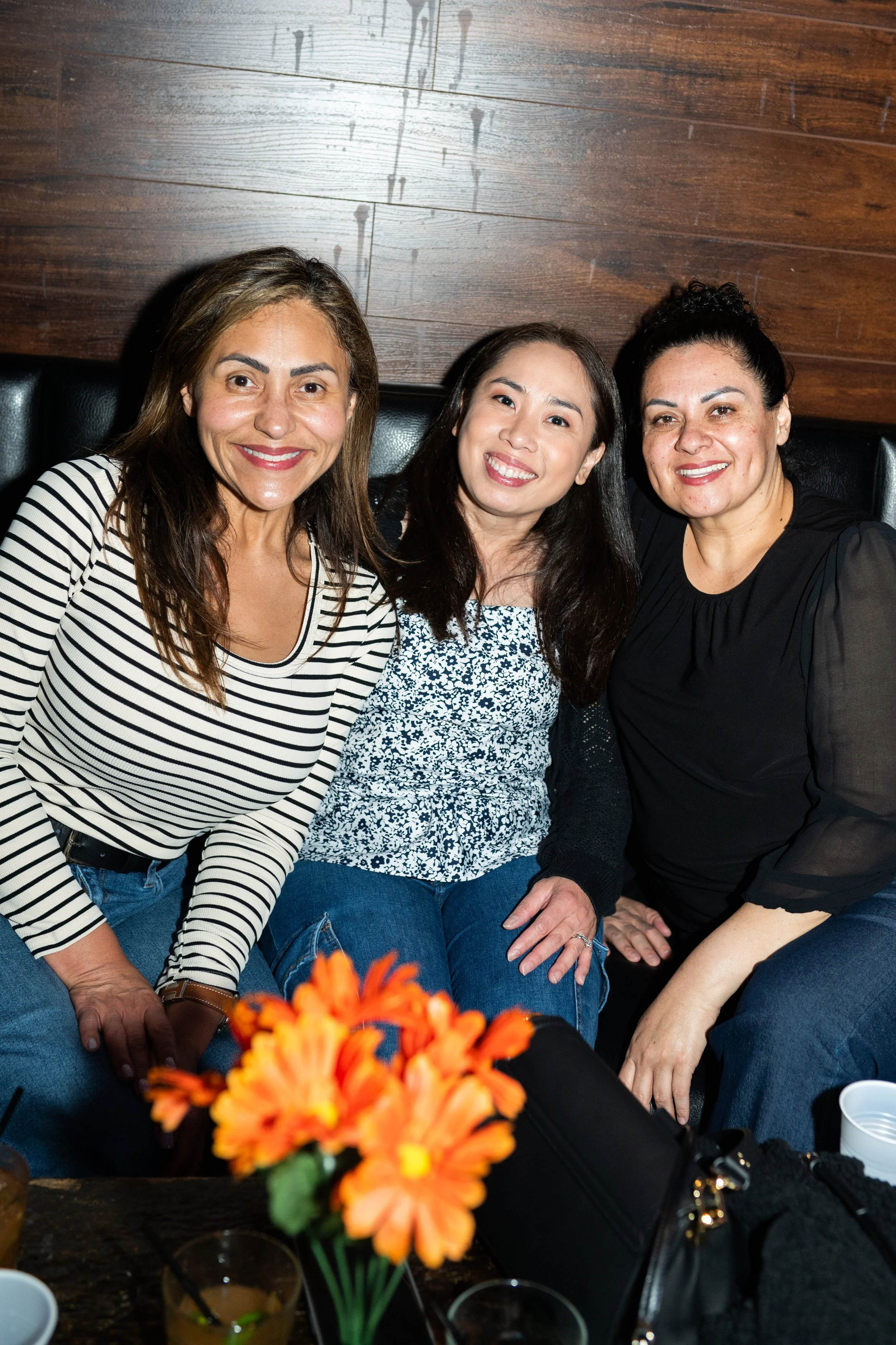 Three women sitting close together on a bench, smiling at the camera, in a dimly lit restaurant or bar with a wooden wall behind them. There is a table in front with orange flowers, drinks, and a black handbag.