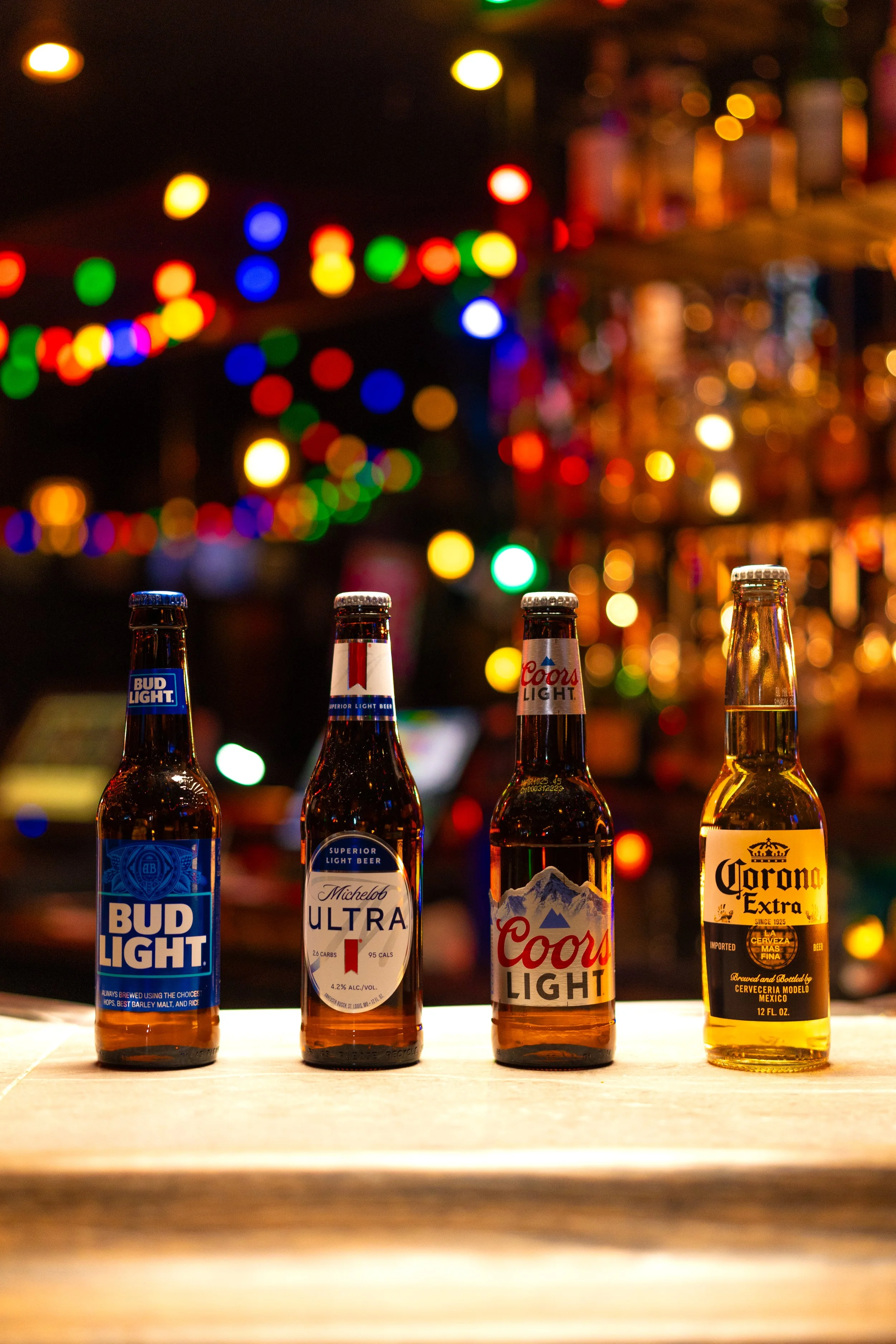 Four bottles of beer and lighted colorful string lights in the background on a bar counter.