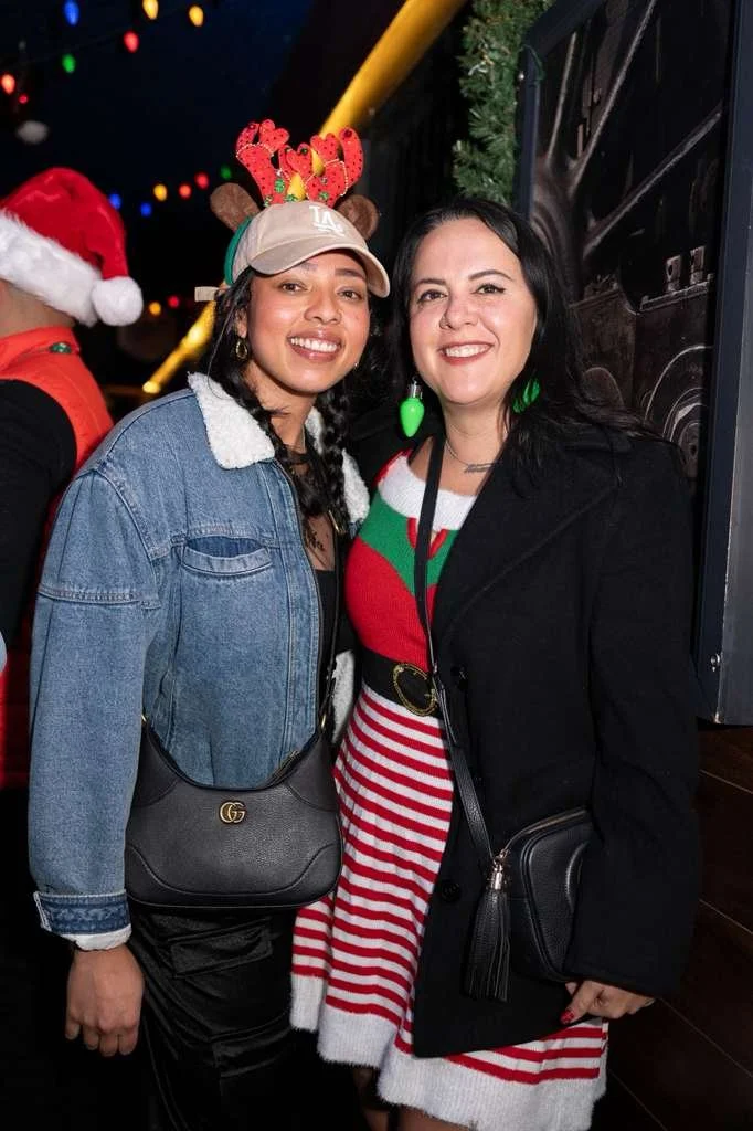 Two women smiling at a Christmas party, one wearing reindeer antlers and a Santa hat, the other dressed in a striped Christmas sweater and festive earrings.
