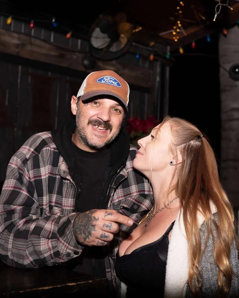 Two people, a man with a beard and tattoos wearing a baseball cap and plaid shirt, and a woman with long red hair wearing a black top and jacket, are close together, smiling and looking at each other in a cozy, festively decorated indoor setting.