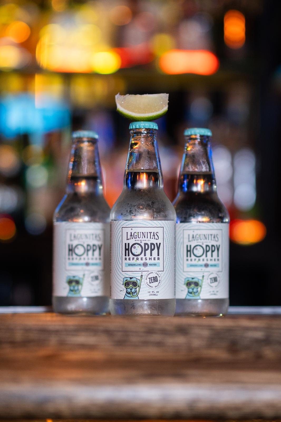 Three glass bottles of Lagunitas Hoppy Refresher sparkling hop water with a lime wedge balanced on the middle bottle's cap, on a wooden surface with a colorful blurred bar background.