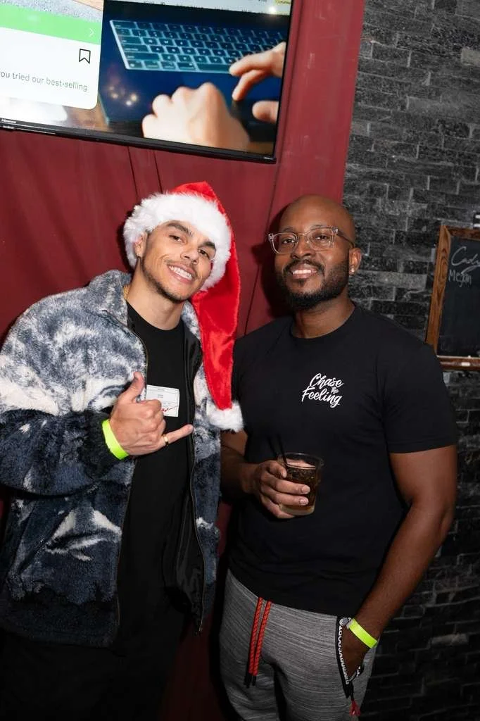 Two men standing together at a social event, one wearing a Santa hat and a patterned jacket, the other in a black t-shirt with 'Chase Feeling' written on it, holding a drink, smiling. A TV screen showing a person typing on a laptop is in the backgrou