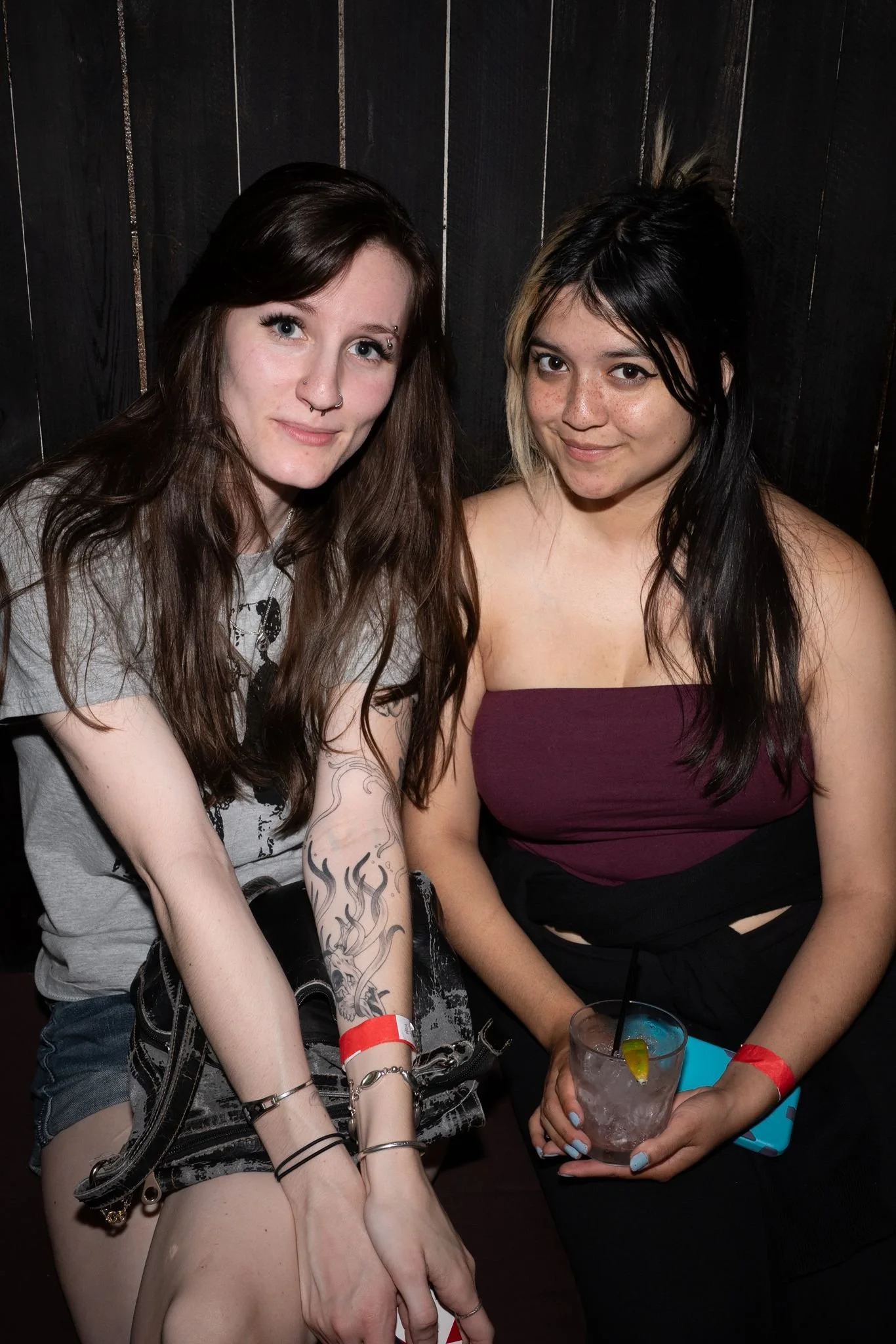 Two young women sitting together in a bar or club, one with brown hair and tattoos on her arm, and the other with dark hair and a beverage in her hand, both smiling at the camera.