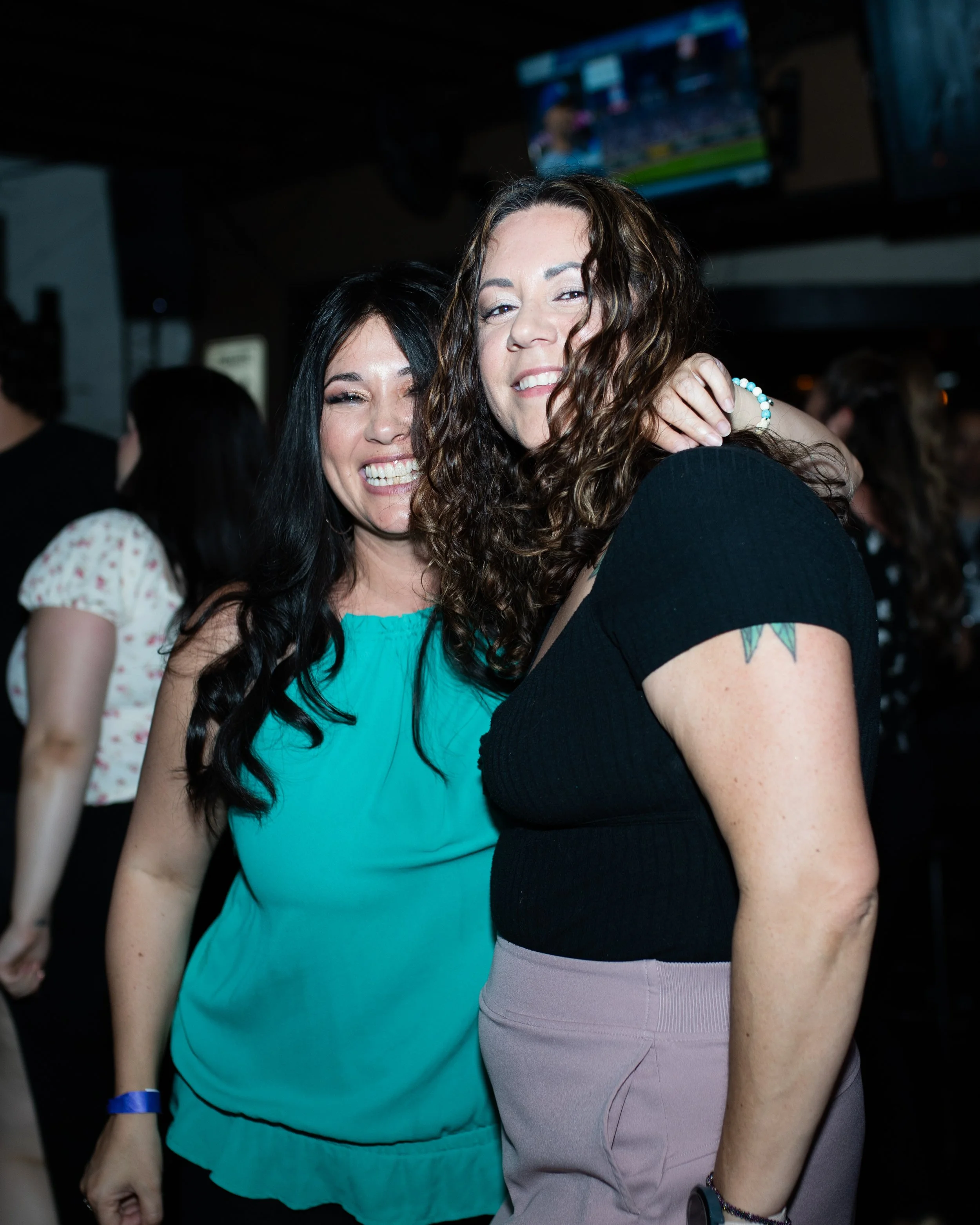 Two women smiling and posing together in a crowded indoor setting, possibly a bar or club, with a television screen visible in the background.
